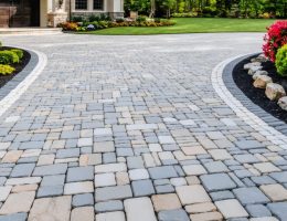 Luxurious driveway design featuring a mix of granite cobblestones, limestone borders, and travertine accents surrounded by a landscaped front yard.