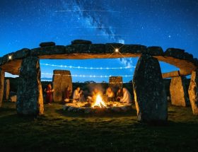 Artistic depiction of a megalithic stone circle with celestial alignments and ghostly figures in traditional clothing celebrating, symbolizing the connection between ancient and modern stone festivals.