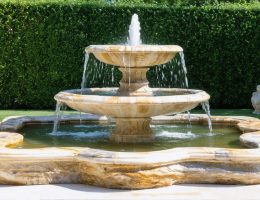 A large, hand-carved natural stone fountain with tiered basins, made from limestone, granite, and marble, nestled within a lush, landscaped garden. The fountain features flowing water and showcases the intricate veining and color variations of the stone.