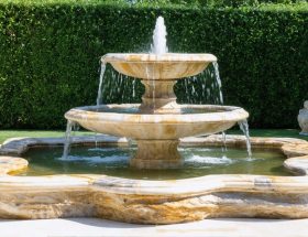 A large, hand-carved natural stone fountain with tiered basins, made from limestone, granite, and marble, nestled within a lush, landscaped garden. The fountain features flowing water and showcases the intricate veining and color variations of the stone.