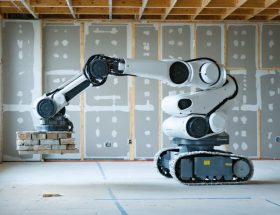 Drywall-finishing robot applying joint compound on a commercial jobsite, with a technician observing and blurred stone slabs on A-frames in the background, illustrating parallels between drywall automation and stone installation.