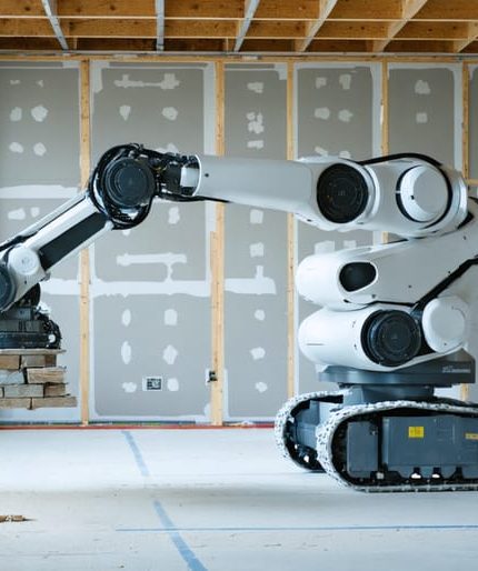 Drywall-finishing robot applying joint compound on a commercial jobsite, with a technician observing and blurred stone slabs on A-frames in the background, illustrating parallels between drywall automation and stone installation.