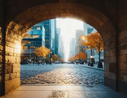 Low-angle wide photo of a weathered stone archway and cobblestone street leading toward glass skyscrapers at golden hour, with street trees and pedestrians softened by atmospheric haze.