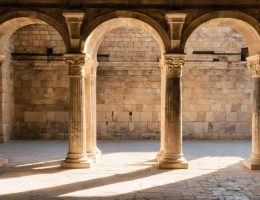 Reused Roman marble columns with mismatched capitals in a later stone church colonnade, warmly side-lit, with softly blurred arches and masonry in the background.