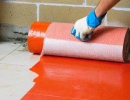 Hand using a paint roller to apply red waterproofing membrane to a cement-board shower corner and curb with mesh tape, with a blurred drain, trowel, and stacked marble tiles in the background.