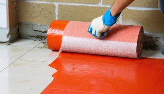 Hand using a paint roller to apply red waterproofing membrane to a cement-board shower corner and curb with mesh tape, with a blurred drain, trowel, and stacked marble tiles in the background.