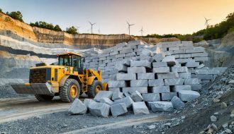 Wide view of a modern granite quarry with cut stone blocks and a wheel loader under golden hour side light, stepped quarry walls, dust mist, and distant wind turbines in the background.