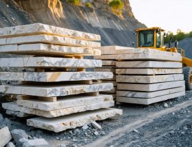 Stacked limestone and marble slabs in a quarry yard at golden hour, detailed textures in the foreground with a blurred quarry wall, wheel loader, and short-haul truck in the background.
