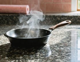 Close-up of a granite countertop holding a steaming cast-iron skillet, with a blurred roll of pink fiberglass insulation in the background under soft natural light.