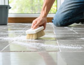 Hand scrubbing white chalky efflorescence from natural stone tiles and grout lines with a soft-bristle brush in soft daylight; bucket and sponge blurred in the background.