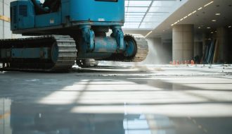 Robotic arm with a vacuum lifter placing a large natural stone slab on a lobby floor, with stacked slabs and glass walls visible in a softly blurred background.