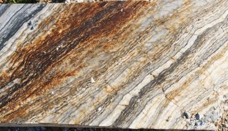 Close-up of a granite surface with rust-colored oxidation streaks, fine pitting, and exposed quartz and feldspar crystals, side-lit by warm sunlight, with a softly blurred outdoor background.