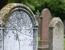 Close-up of a deteriorating marble gravestone showing lichen growth, black crust from pollution, and fine cracks, photographed in soft overcast light with blurred headstones and trees in the background.