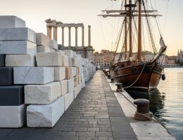 Stacks of marble, granite, and limestone on an old European quayside beside a wooden cargo ship at golden hour, with a softly blurred skyline suggesting Roman columns and a Gothic cathedral spire.