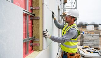Mason installing a limestone rainscreen panel onto thermally broken stainless steel brackets over mineral wool continuous insulation and a red fluid-applied air barrier, with scaffolding and stacked stone softly blurred in the background