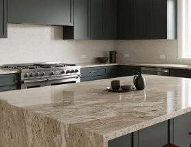 Contemporary kitchen with a large warm neutral granite island slab showing an eased edge, matte charcoal cabinets, stainless range, and a subtle engineered tile backsplash under soft natural daylight.