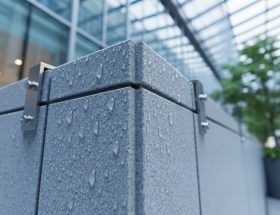 Low-angle close-up of thin granite facade panels with water droplets beading on the surface, showing hydrophobic treatment and precise mounting; blurred glass atrium, greenery, and pedestrians in the background