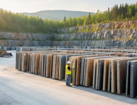 Engineer in safety gear examining stacked natural stone slabs at a clean quarry in golden hour side light, with reforested benches, rolling hills, and a compact wheel loader in the background.