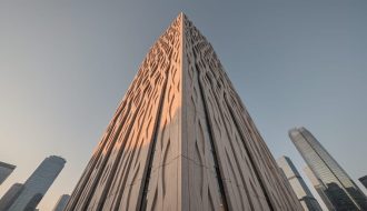 Low-angle view of a contemporary skyscraper clad in thin natural-stone rainscreen panels with CNC-carved relief, lit by golden-hour side light, with distant glass towers and a blue-gray sky in the background.
