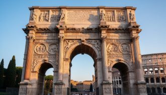 Low-angle view of the Arch of Constantine showing mismatched reused marble relief panels and columns in warm late-day light, with the Colosseum softly blurred behind.