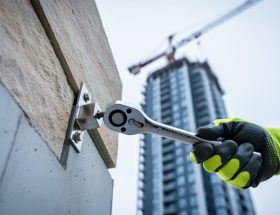 Gloved engineer tightens a stainless steel anchor bracket securing a stone cladding panel to concrete, low-angle view with a blurred high-rise facade and crane in the background under soft overcast light