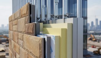 Cutaway view of a natural stone facade showing concealed clips, ventilated rainscreen cavity, continuous rigid insulation, and air/water barrier on a modern building, with a softly blurred construction site and skyline in the background.