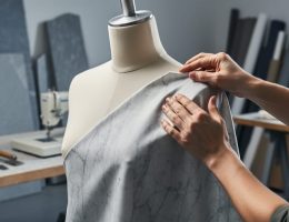 Close-up of hands applying an ultra-thin marble veneer to a mannequin in a fashion studio, with chiseling tools, stone slabs, and fabric rolls softly blurred in the background.