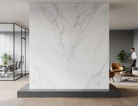 Freestanding floor-to-ceiling veined white marble wall with a granite base defining zones in a contemporary open-plan office, with soft natural light, glass meeting room, collaborative lounge, indoor plants, and a few motion-blurred employees in the background.