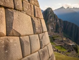 Close-up of interlocking polygonal granite stones in a Machu Picchu wall under warm side light, showing seamless mortarless joints, with softly blurred terraces and distant Andean mountains in the background.