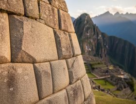 Close-up of interlocking polygonal granite stones in a Machu Picchu wall under warm side light, showing seamless mortarless joints, with softly blurred terraces and distant Andean mountains in the background.