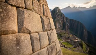 Close-up of interlocking polygonal granite stones in a Machu Picchu wall under warm side light, showing seamless mortarless joints, with softly blurred terraces and distant Andean mountains in the background.