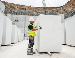 Quarry technician in safety gear inspects a large white marble block, hand tracing veining and checking for fissures, with stacked blocks, quarry face, and a wire saw visible under bright overcast daylight.