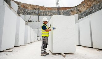 Quarry technician in safety gear inspects a large white marble block, hand tracing veining and checking for fissures, with stacked blocks, quarry face, and a wire saw visible under bright overcast daylight.