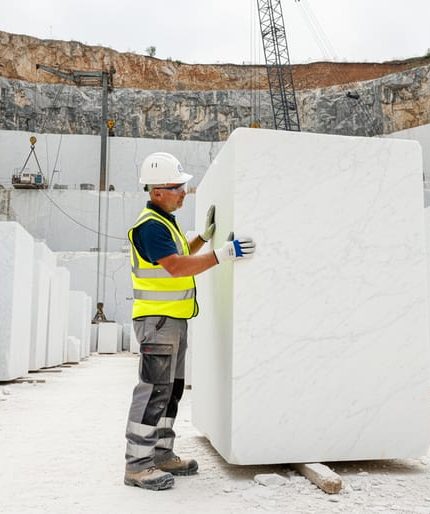 Quarry technician in safety gear inspects a large white marble block, hand tracing veining and checking for fissures, with stacked blocks, quarry face, and a wire saw visible under bright overcast daylight.