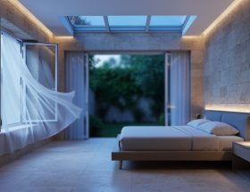 Modern bedroom with limestone wall and travertine floor, open windows and flowing sheer curtains in cool pre-dawn light, suggesting natural cross-ventilation with a leafy courtyard visible outside.