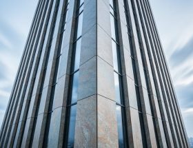 Low-angle photo of a modern high-rise with natural stone cladding at the corner, sharp panel joints and texture visible, with fast-moving clouds and a distant skyline in the background.