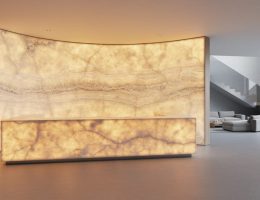 Curved backlit onyx feature wall with matching countertop glowing amber, veining clearly visible, photographed at eye level in a modern interior with minimal seating and a staircase in the background.
