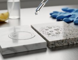 Close-up of marble and granite samples on a stainless workbench as a glass dropper applies liquid; marble shows slight etching while granite beads droplets, with a lemon wedge and unlabeled spray bottle softly blurred in the background.