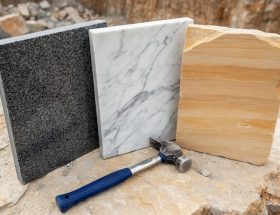 Close-up three-quarter overhead view of polished granite, white-veined marble, and warm sandstone slabs on a rough stone surface next to a geologist’s hammer, with a quarry wall softly blurred in the background under diffused daylight.
