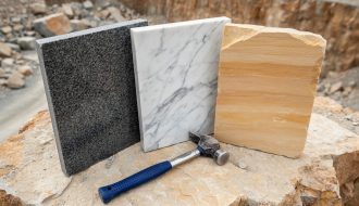 Close-up three-quarter overhead view of polished granite, white-veined marble, and warm sandstone slabs on a rough stone surface next to a geologist’s hammer, with a quarry wall softly blurred in the background under diffused daylight.