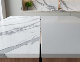 Two large slabs of Calacatta marble and light gray quartz displayed side by side on a modern kitchen island, with soft daylight and a blurred background of cabinetry, a brass faucet, and a small potted herb.