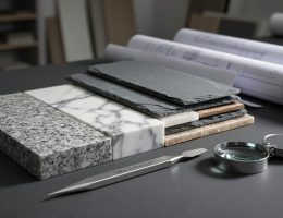 Close-up angled view of granite, marble, slate, and quartzite samples on a dark workbench with a metal scratch pick and hand lens, side-lit to emphasize texture and luster, with a blurred architecture studio and rolled plans in the background.