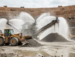Active stone quarry with mist cannons spraying at a crusher and conveyor to control dust, wheel loader operating in foreground, terraced rock walls and haul trucks in the background under overcast light.