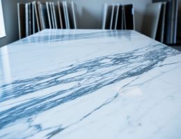 Close-up of polished white marble with natural blue-gray and charcoal veins, shot at a 45-degree angle in soft side daylight, with a blurred stone showroom and stacked slabs in the background.