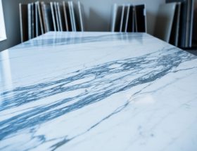 Close-up of polished white marble with natural blue-gray and charcoal veins, shot at a 45-degree angle in soft side daylight, with a blurred stone showroom and stacked slabs in the background.
