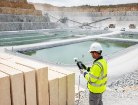 Engineer in a high-visibility vest checks an air monitoring device beside lined settling ponds and stacked limestone blocks at an active quarry, with terraced rock walls, haul trucks, and conveyors in the background under bright overcast light.