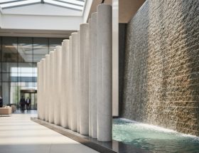Kinetic stone installation in a modern corporate lobby with rotating polished marble columns and a shimmering textured stone water wall, photographed at eye level with soft daylight and blurred visitors in the background.