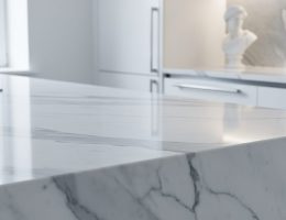 Close-up of a polished white Carrara marble kitchen countertop with gray veining, side-lit to show subtle translucency, with a softly blurred modern kitchen and a small classical marble bust in the background.