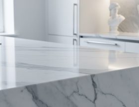 Close-up of a polished white Carrara marble kitchen countertop with gray veining, side-lit to show subtle translucency, with a softly blurred modern kitchen and a small classical marble bust in the background.