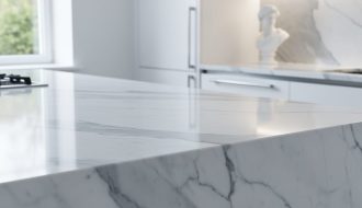 Close-up of a polished white Carrara marble kitchen countertop with gray veining, side-lit to show subtle translucency, with a softly blurred modern kitchen and a small classical marble bust in the background.